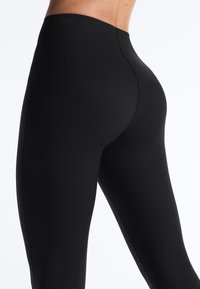 Woman wearing black high-waist leggings, shown from the side and back, highlighting the smooth fabric and fitted design.