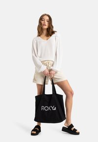 Černá plátěná taška s bílým logem "ROXY", spárovaná s bílou texturovanou blůzkou a béžovými šortkami. Modelka má na sobě černé nazouváky.
