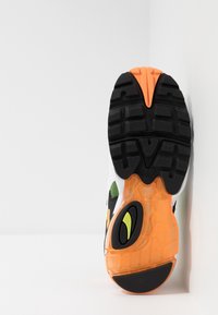 Vit idrottssko med svarta, gröna, gula och orange accentuerade detaljer. Har en texturerad gummisula med djupa mönster och transparenta partier.