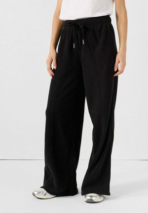 Pantaloni da ginnastica neri a gamba larga con vita regolabile, cuciture laterali e texture morbida, abbinati a scarpe sportive argento.