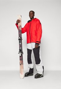 Punane veekindel suusajakk, millel on tõmblukk, koos mustade ja valgete suusapükstega. Isik hoiab mustrilist snowboardi, millel on sidemed.