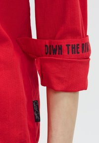 Rotes, strukturiertes Hemd mit hochgerollten Ärmeln, versehen mit schwarzem, gesticktem Text "DOWN THE RIVER" und einem kleinen Logosticker an der Seite.