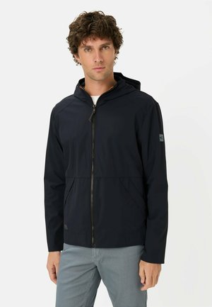 Mężczyzna z kręconymi brązowymi włosami, ubrany w czarną bluzę z kapturem na zamek i jasnoszare spodnie, stojący na jednolitym białym tle.