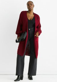 Burgundy knit cardigan with long sleeves, open front, and two pockets, paired with a black top and high-waisted black jeans. Black heels.