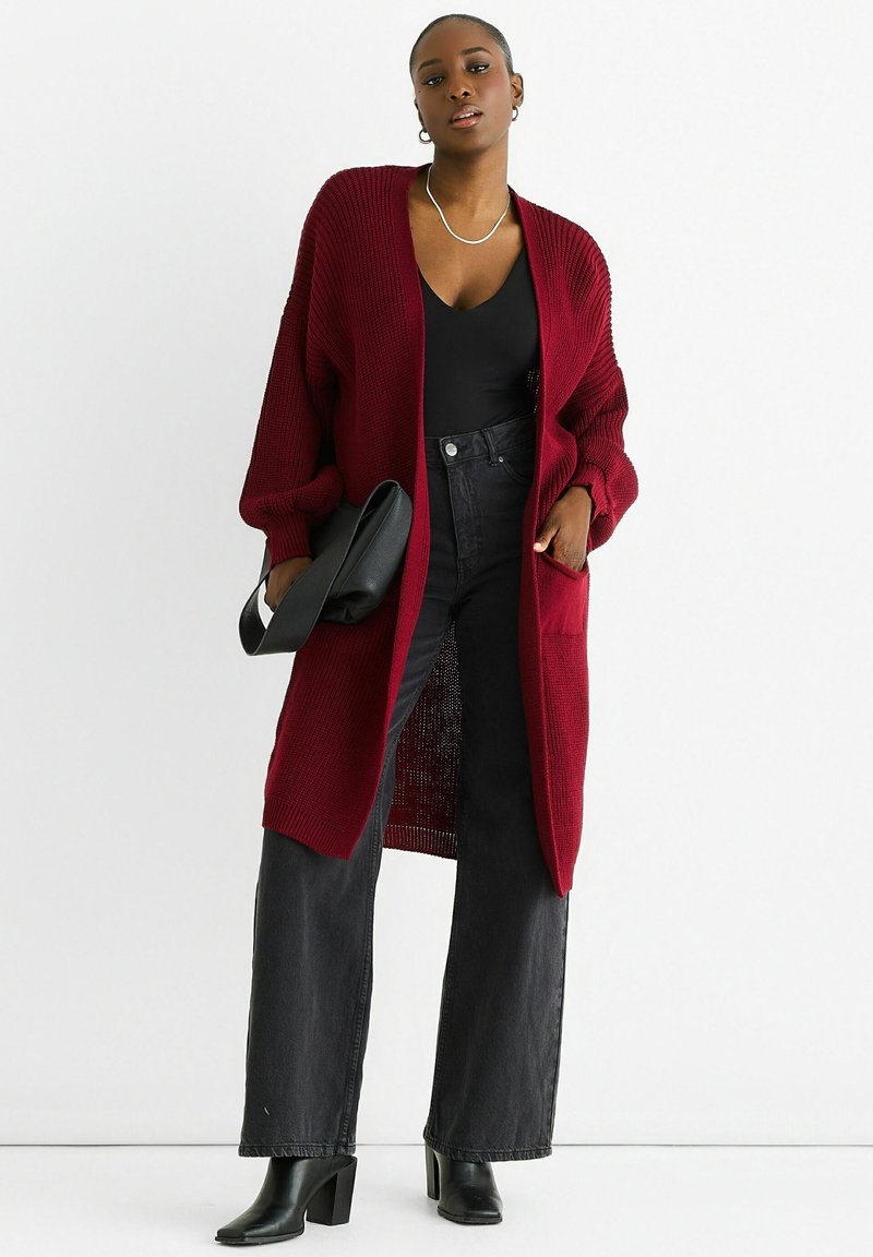 Burgundy knit cardigan with long sleeves, open front, and two pockets, paired with a black top and high-waisted black jeans. Black heels.