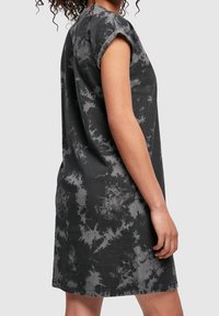 Femme portant une robe tie-dye à manches courtes noire et grise, vue de profil avec le bras détendu le long du corps sur un fond uni.