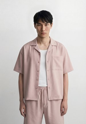 Homme portant une chemise à manches courtes rose pâle boutonnée et un pantalon assorti avec un débardeur blanc, se tenant devant un fond gris uni.