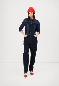 Denim kombinezon temno modre barve z gumbom spredaj in dolgimi rokavi. V kombinaciji s rdečo pleteno kapo in rdeče-bele čevlje s peto.