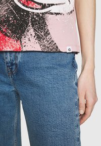 La mano de una persona descansa en la cadera, vistiendo una camiseta gráfica rosa con salpicaduras de rojo y negro, combinada con jeans de mezclilla azul.