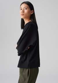 Schwarzes, übergroßes Sweatshirt mit langen Ärmeln, rundem Ausschnitt und weicher Textur, kombiniert mit olivgrünen, plissierten Hosen.