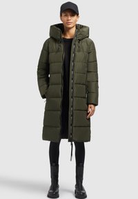 Abrigo acolchado largo de color verde oliva con capucha, diseño de acolchado, cremallera frontal y cordón ajustable. Lleva puesto un top negro y lo acompaña con botas negras.