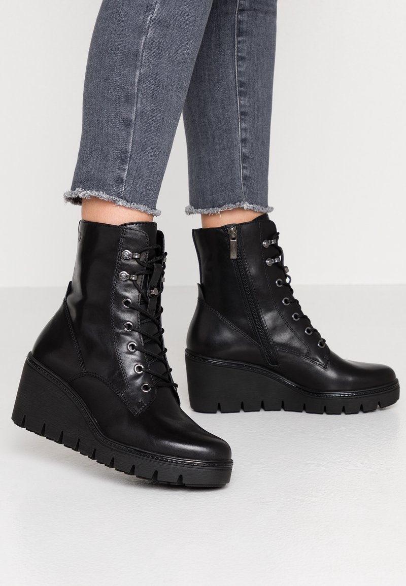 Bottines compensées en cuir noir avec lacets à l'avant et fermeture éclair latérale, portées avec un jean gris court à ourlet effiloché sur fond blanc.