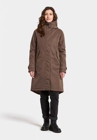 Trench-coat imperméable marron avec col montant, poches avant et dos légèrement plus long, porté avec un pantalon sombre et des bottes noires.