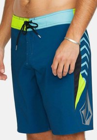 Bañador azul con un acento negro y amarillo neón. Presenta una cintura con cordón y un patrón en zigzag en el lateral. Textura de tela suave.