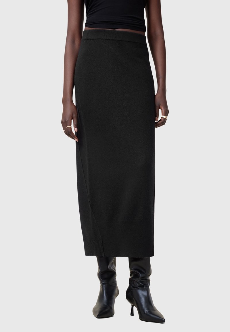 Femme portant une jupe midi noire ajustée, un haut noir sans manches, des bottines noires à talons pointues, et des bagues dorées aux deux mains.