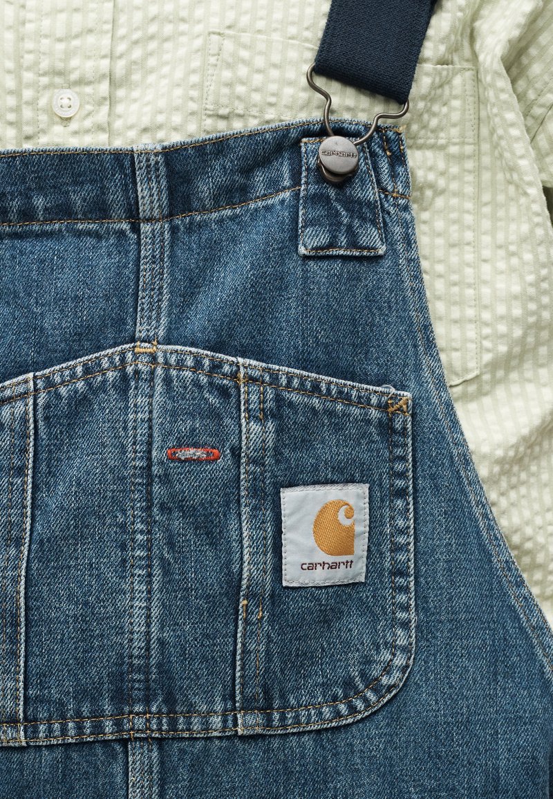 Denim-Carhartt-Latzhose über einem strukturierten hellbeigen Hemd mit Knöpfen, zeigt die Fronttasche und den verstellbaren Riemenverschluss.