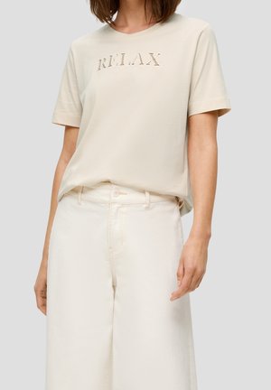 Kobieta w kremowym t-shircie z krótkim rękawem z napisem „RELAX” i kremowych szerokich spodniach z wysokim stanem na jednolitym tle.