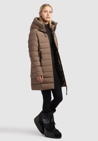 Brown puffer coat with a high collar, zippered front, and side pockets. Worn with black leggings and chunky black snow boots.