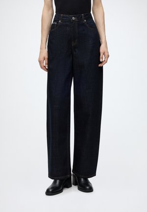 Personne portant un jean large bleu foncé et des bottines noires à talons, debout, les mains détendues le long du corps, sur un fond uni.