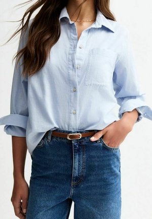 Kvinde iført lyseblå skjorte med knapper og opkørte ærmer, stoppet ned i højtaljede blå jeans med brunt bælte.