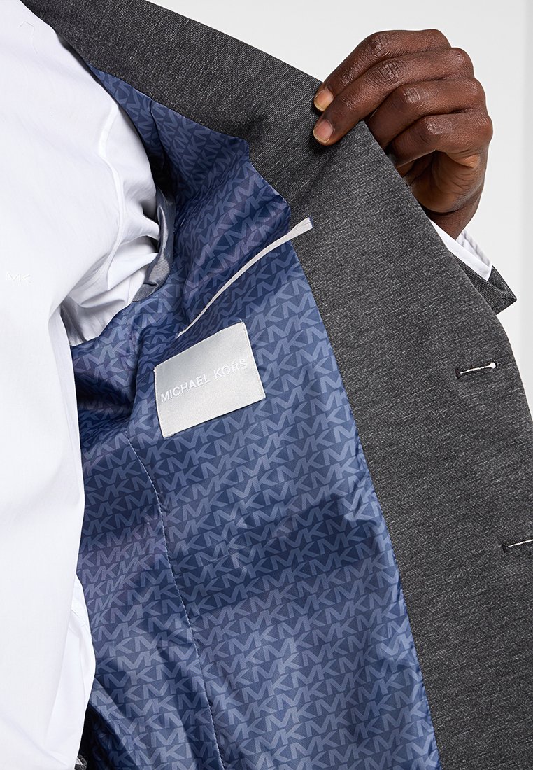 Mano sosteniendo un traje gris abierto que muestra el forro azul con un patrón repetido del logo MK y una etiqueta blanca con "Michael Kors" en el interior.