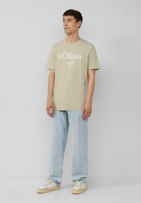 Lichtbeige katoenen t-shirt met grote witte bedrukte merknaam en slogan. Gepaard met lichtblauwe recht gesneden jeans en casual sneakers.