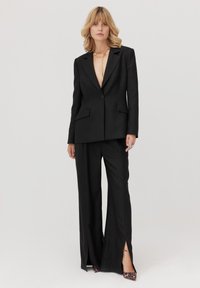 Czarny, dopasowany garnitur z marynarką o głębokim dekolcie w serek i szerokimi spodniami z bocznymi rozcięciami. Dopełniony złotą biżuterią i szpilkami w zwierzęcy wzór.