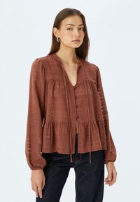Blusa color ruggine realizzata in tessuto leggero, con chiusura frontale a bottoni, design peplo, maniche lunghe e sottili righe testurizzate.