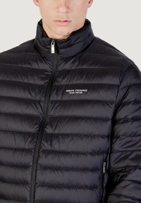 Chaqueta acolchada negra con un alto cuello, cierre de cremallera y diseño de acolchado horizontal. Presenta un logo en el lado izquierdo del pecho y un bolsillo lateral.