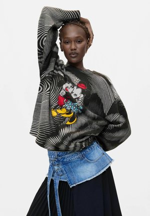 Femme aux cheveux tressés posant avec un pull noir et gris à motifs Mickey et Minnie Mouse et une jupe en denim noire superposée.