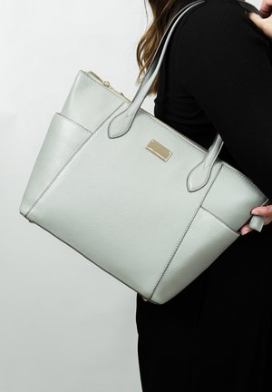 Light green leather tote bag with structured shape, dual handles, and a gold-tone plaque detail. Zippered top and side pocket visible.