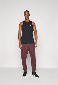 Zwart mouwloos sporttop met wit logo, bordeaux joggingsbroek met zwarte zijkanten, en zwarte sportschoenen.