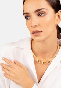 Collarín de oro liso y redondeado con múltiples colgantes en forma de lágrima colgando por debajo. La modelo lleva una camisa blanca.