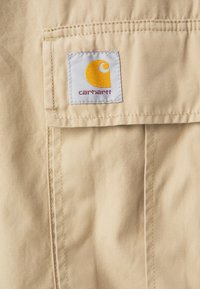 Nærbillede af beige stof med en syet lomme og et mærke med et cirkulært logo i gult og lilla, mærket "Carhartt."