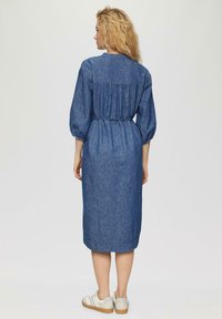 Dunkelblaues Denim-Midikleid mit Puffärmeln, einem gerafften Rücken und einem Bindegürtel in der Taille. Kombiniert mit weißen Sneakers.