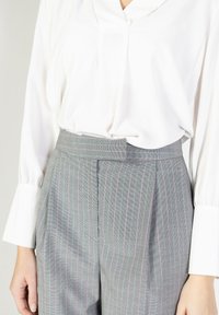 Pantaloni grigi a righe con dettagli in vita e pieghe, abbinati a una blusa bianca a maniche lunghe con scollatura a V.