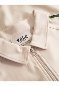 Cream zip-up jacket featuring a fold-down collar, branded label stitched on fabric, and smooth, textured material.