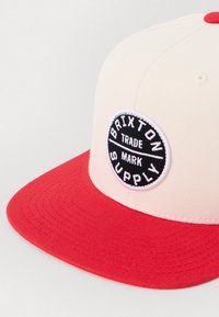 Căciulă roșie și crem cu brâu plat, având o aplicație circulară neagră pe care scrie "BRIXTON SUPPLY." Brâul are o textură vizibilă cu cusături.