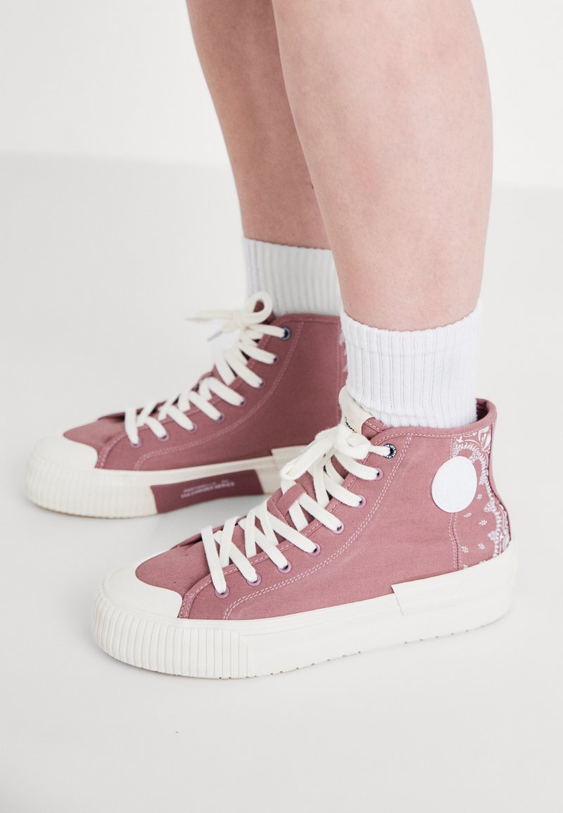 Zapatillas altas de lienzo rosa con suelas y acentos de goma blanca. Presentan cordones blancos, textura acanalada y costura decorativa en el lateral.