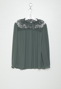 Blouse à manches longues vert foncé avec un empiècement en dentelle, taille froncée et tissu lisse. Caractéristiques : coupe fluide et poignets à boutons. Accrochée à un crochet.