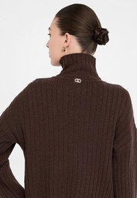 Maglione marrone a coste con colletto alto, caratterizzato da un piccolo logo sul retro. La trama è spessa, aumentando il calore e il comfort.