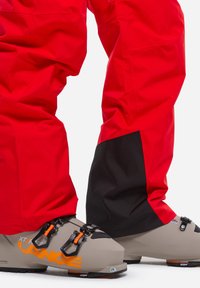 Red ski pants with a black lower section, paired with grey ski boots featuring orange accents and black hardware. Durable fabric with smooth texture.