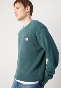 Teal trui met een gestructureerde brei, ronde halslijn en geribbelde manchetten. Heeft een klein logo-opdruk op de voorkant. Gepaard met blauwe jeans.