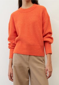 Ljus orange stickad tröja med ribbat mönster, vid rund halsringning och lösa ärmar. Matchad med ljusbeige byxor.