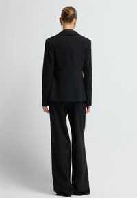 Zwart kostuum met een getailleerde blazer met schoudervullingen en rechte broekspijpen. De stof heeft een subtiel streepjespatroon.