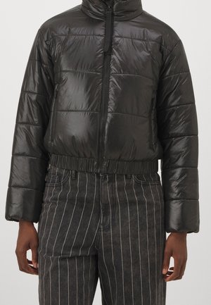 Doudoune noire brillante matelassée avec fermeture éclair sur le devant, portée avec un pantalon gris foncé à fines rayures sur une personne devant un fond uni.