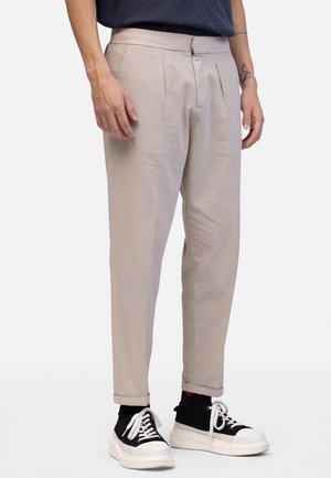 Mand iført lyse beige plisserede bukser, sorte sokker og sort-hvide platformsneakers, står foran en ensfarvet baggrund.