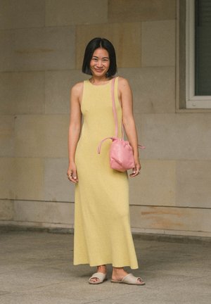 Une femme se tient à l'intérieur, portant une robe maxi jaune sans manches, des sandales beiges sans lacets, et un sac à bandoulière rose à cordon.