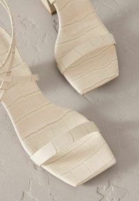 Sandales à bride couleur crème avec une texture crocodile, forme à bout carré et petit talon carré, dotées de fines brides sur le pied.