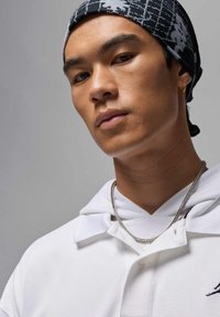 Biała koszulka polo z kapturem, teksturowana tkanina i srebrny naszyjnik z łańcuszkiem. Czarna, wzorzysta bandana noszona na głowie, zbliżenie twarzy.